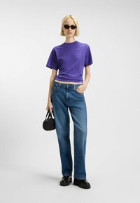 Purple short-sleeve t-shirt with a round neckline, paired with light blue straight-leg jeans. Holding a small black handbag, wearing sunglasses.