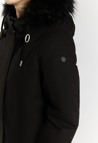 Sort parka med en faux fur hætte, lynlåslukning foran og sølvfarvede hardware-detaljer. Har et logo-badge på ærmet og en tekstureret stofoverflade.