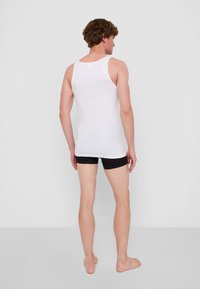 Vit ärmlös linne i bomull, figurnära design. Bärs med svarta shorts. Utsikten visar modellen bakifrån och benen.