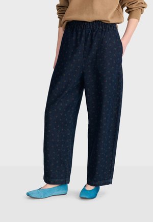Personne portant un pantalon ample bleu foncé avec de petits motifs rouges, un haut marron à manches longues et des chaussures plates bleu vif, debout les mains dans les poches.
