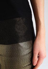 Top negro con una sección inferior metálica dorada texturizada. Incluye una tela suave y ligera con un corte ajustado. Detalle de costura visible.