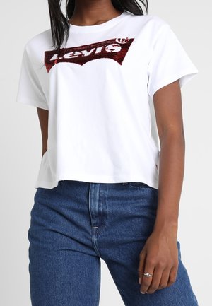 Biała bawełniana koszulka o skróconym kroju z czerwoną, cekinową stroną logo Levi's na piersi. Połączona z niebieskimi dżinsami.