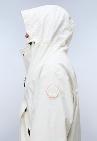 Άτομο σε προφίλ που φοράει λευκό μπουφάν με κουκούλα και κυκλικό σήμα με το λογότυπο Napapijri στο πάνω μέρος του μπράτσου, πάνω σε μονόχρωμο φόντο.