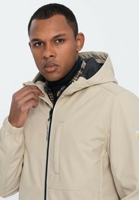 Beige Kapuzenjacke mit elastischen Bündchen, zentriertem Reißverschluss und Seitentasche. Über einem schwarzen Rollkragenpullover und einem karierten Hemd getragen. Glatte Stofftextur.