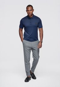 Granatowa koszulka polo z kołnierzykiem i trzema guzikami, zestawiona z jasnoszarymi, dopasowanymi spodniami i czarnymi butami. Na koszulce znajduje się logo.