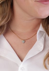 Gouden ketting met een lichtblauwe hartpendant, gedragen over een witte blouse met kraag. De ketting heeft ovale schakels en een glanzende afwerking.