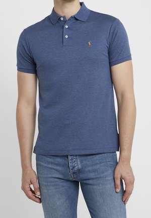 Mann trägt ein blaues Poloshirt mit kurzen Ärmeln und drei Knöpfen sowie hellblaue Jeans vor einfarbigem Hintergrund.