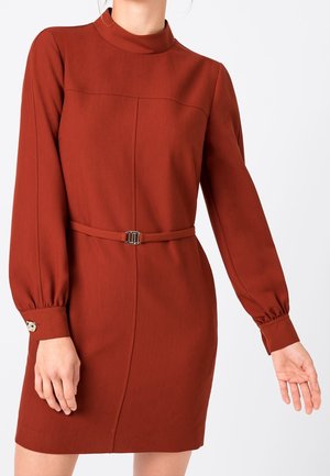 Vrouw die een roestrood jurkje met lange mouwen draagt, met een hoge halslijn, een dunne riem in de taille en met knopen afgewerkte manchetten.