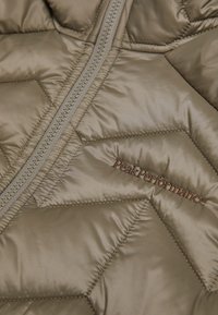 Beige pufferjack met chevronstiksel, een rits aan de voorkant en geborduurd 'PeakPerformance' logo in een contrasterende kleur.