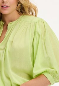 Blusa verde chiaro con scollatura arricciata, maniche a sbuffo e chiusura con bottoni. Tessuto trasparente e liscio con leggera texture e ampiezza.