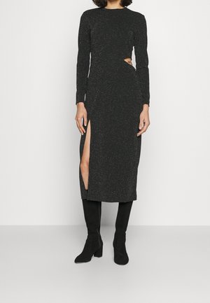 Kobieta w czarnej, długorękawowej sukience midi z bocznym wycięciem i rozcięciem sięgającym uda, w połączeniu z czarnymi, sięgającymi kolan, wysokimi butami na obcasie.