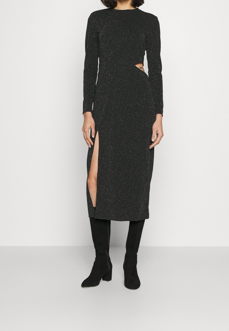 Femme portant une robe midi noire à manches longues avec une découpe sur le côté et une fente haute sur la cuisse, associée à des bottes noires à talons montant jusqu'aux genoux.
