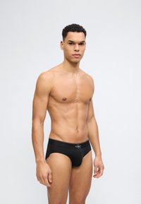 Μυώδης άνδρας στέκεται, φορώντας μαύρα εσώρουχα Calvin Klein, με κοντά σγουρά μαλλιά, ουδέτερη έκφραση, σε απλό ανοιχτό γκρι φόντο.