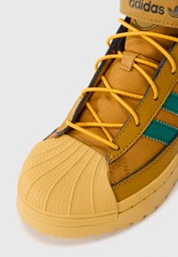 Sneaker amarelo-dourado com biqueira de borracha texturizada, parte superior em malha, riscas turquesa e atacadores laranja em contraste. Apresenta uma sola robusta.