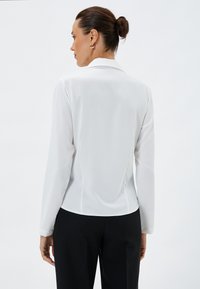 Hvid langærmet bluse med krave, glat tekstur, tætsiddende design og synlige sømme, parret med sorte bukser.