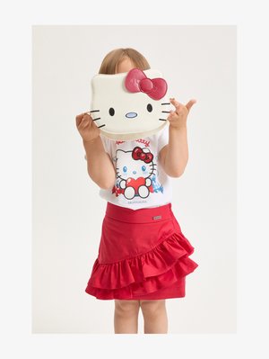Niño con falda roja con volantes y camiseta de Hello Kitty sostiene un bolso con la cara de Hello Kitty frente a su rostro, con un fondo liso.