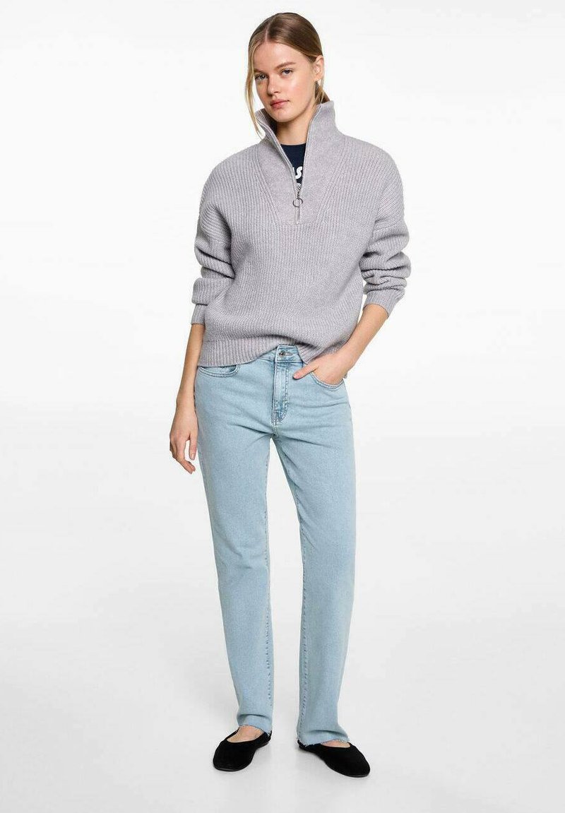 Svetlosivý rebrovaný pulóver s polovičným zipsom na golieri, skombinovaný so svetlomodrými džínsami s rovnými nohavicami a čiernymi šľapkami. Celkový vzhľad je hladký.