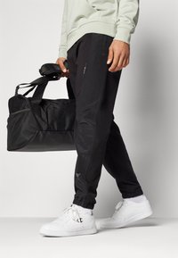 Sorte joggebukser med teksturerede sidepaneler, elastiske manchetter og lynlåslommer, parret med hvide sneakers og en sort duffeltaske.