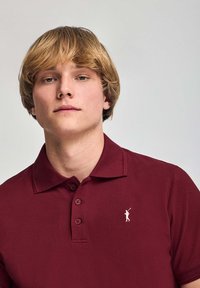 Burgundy pikétröja i texturerad tyg med en treknappling, korta ärmar och en liten vit golfspelare-logotyp på bröstet.