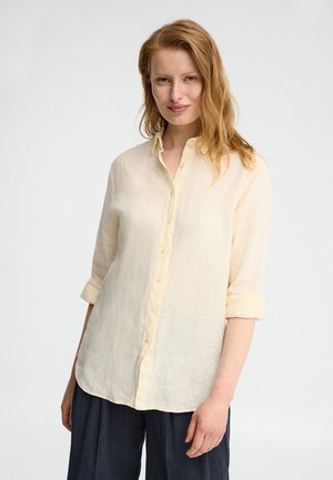 Kobieta o jasnoczerwonych włosach ubrana w kremową koszulę zapinaną na guziki z podwiniętymi rękawami oraz ciemne, luźne spodnie, stojąca na jednolitym tle.