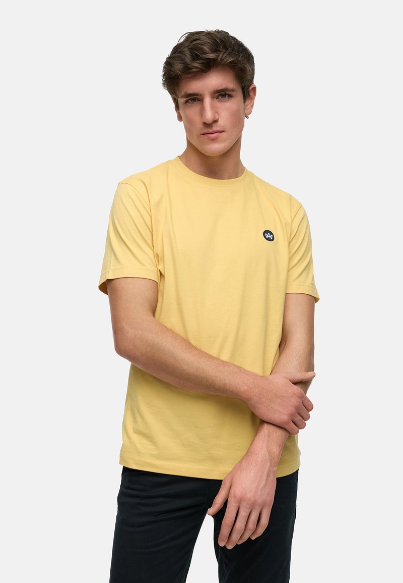Een gele katoenen t-shirt met een ronde halslijn en korte mouwen, voorzien van een klein zwart logo op de linkerkant van de borst. Gestyled met donkere broeken.