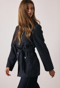 Ciemnogranatowy płaszcz z teksturowanej tkaniny, z paskiem do wiązania w talii, szerokimi rękawami i ozdobnymi guzikami. Casualowy design idealny do warstwowania.