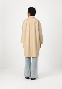 Cappotto trench beige con un fit rilassato, design di lunghezza media e maniche larghe, abbinato a jeans a zampa d'elefante blu chiaro e scarpe nere.