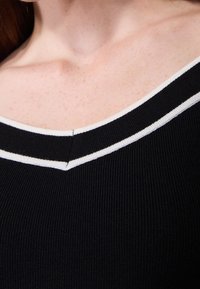 Haut noir côtelé avec un col en V orné d'une bande horizontale blanche le long de l'encolure et une texture subtile.