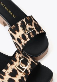 Sandalias deslizantes con estampado de leopardo, con una plantilla sintética negra, tacón grueso y acentos en dorado con el logo. Presenta una correa de tela texturizada.
