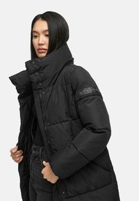 Black puffer jacket with a high collar, button closures, and zippered front. Features quilted texture and a logo on the sleeve.
