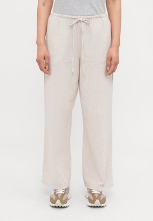 Lyse beige bukser med vid ben og snøre i taljen, båret med beige og hvide sneakers, model stående mod en ensfarvet baggrund.