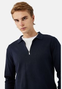 Navyblaues Langarmshirt mit Reißverschlusskragen, aus strukturiertem Stoff. Darunter ist ein weißes Rundhals-T-Shirt sichtbar.