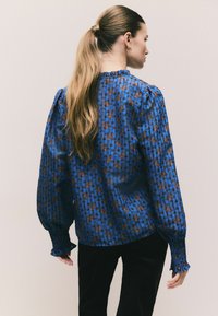 Blusa blu a fantasia con maniche lunghe, a palloncino e polsini arricciati. Presenta un tessuto strutturato con dettagli floreali in tonalità marroni.