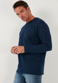 T-shirt a maniche lunghe blu navy con scollatura rotonda, realizzato in morbido tessuto di cotone. Presenta fessure laterali e una vestibilità comoda. Indossato con jeans chiari.