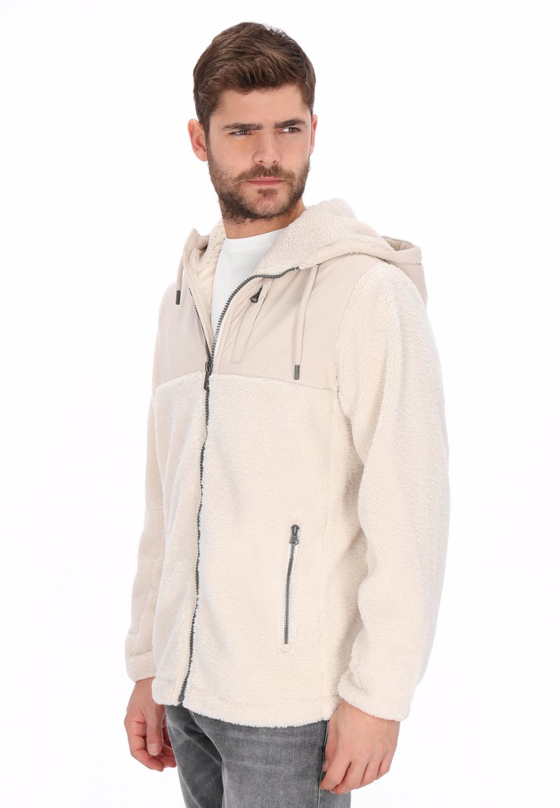 Chaqueta de forro polar en blanco roto con un panel superior beige. Cuenta con capucha, cremallera frontal y bolsillos laterales. Textura suave con secciones de colores contrastantes.
