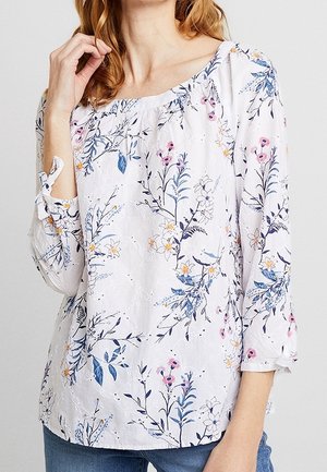 Kvinde iført en hvid bluse med blå, pink og gule blomsterprint og 3/4 ærmer med bindedetaljer, kombineret med blå jeans.