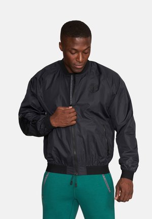 Mężczyzna zapinający czarną kurtkę bombowiec, ubrany w miętowe spodnie ze ściągaczem, patrzący w dół na jasnym, jednolitym tle.