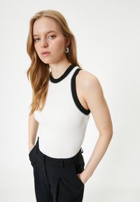Geribbelde witte tanktop met zwarte accenten bij de halslijn en armgaten. Het model draagt zwarte broeken met zakken en staat met de handen in de zakken.