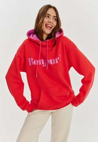 Červená prehnaná mikina s fialovým podšívkou kapucne. Obsahuje text "Bonjour!" v svetlejšej fialovej farbe. Mäkkučkú textúru, klokanie vrecko, elastické manžety.