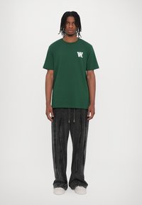 Dark green cotton t-shirt with short sleeves and a white logo on the chest, paired with loose-fitting black denim pants and white sneakers.
