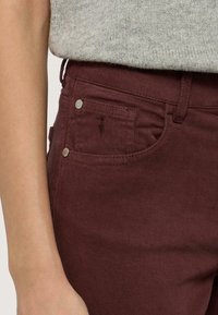 Pantalones de algodón burdeos con un bolsillo frontal, botones metálicos plateados y una textura sutil. La cinturilla cuenta con pasadores para cinturón para un soporte adicional.