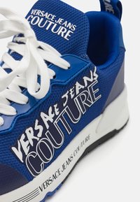 Zapatilla de malla azul con cordones blancos y texto de marca grande en colores contrastantes. Suela intermedia blanca gruesa con suela exterior negra.