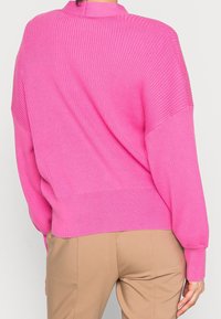 Rosa ribbstickad tröja med hög hals och sänkta axlar. Har långa ärmar och en avslappnad passform. Bärs över beige byxor.