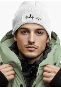White ribbed beanie with a black heartbeat graphic and a runner silhouette on a rectangular patch; worn with a green parka jacket.