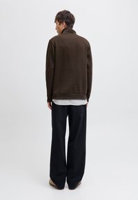 Person iført en brun jakke, stripete skjorte, vide svarte bukser og lysebrune sko, stående vendt bort fra kameraet mot en ensfarget hvit bakgrunn.