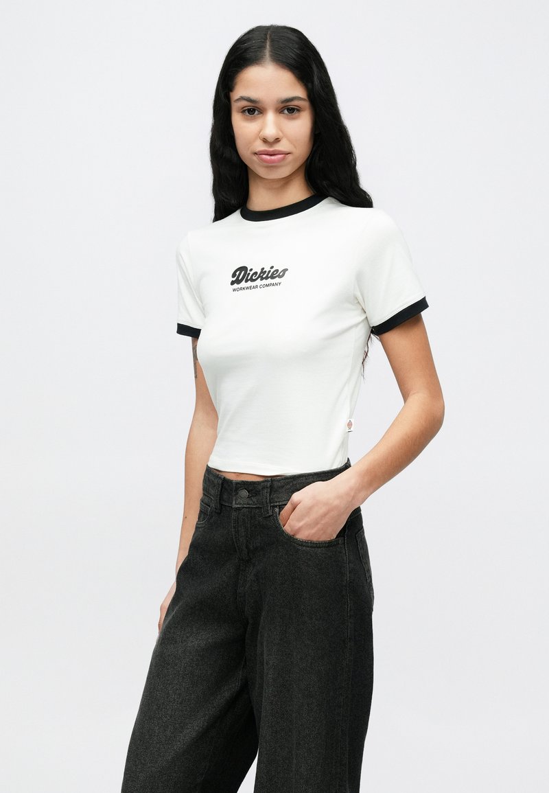 Junge Frau mit langen schwarzen Haaren, die ein weißes Crop-Top mit schwarzem Besatz und schwarze Jeans mit weitem Bein trägt, steht mit einer Hand in der Tasche.