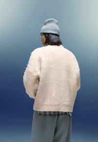 Gruba, kremowa sweter z dzianiny o miękkiej, puszystej teksturze, z ribbowym dołem, noszona na koszuli w kratę i zestawiona z szarymi spodniami.