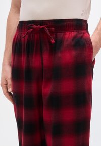 Pantalon de pyjama à carreaux rouges et noirs avec une taille élastique et un cordon de serrage. Comprend des poches latérales et une texture douce et brossée.