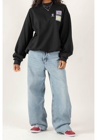 Felpa nera oversized con maniche lunghe, caratterizzata da una grafica colorata sulla tasca del petto, abbinata a jeans baggy blu chiaro e sneaker rosse.