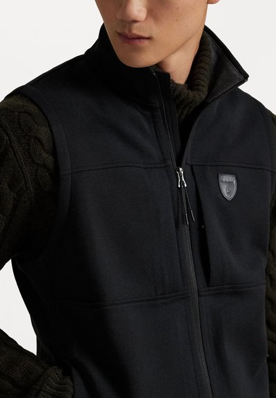 Melnā zip-up veste, kas izgatavota no gluda sintētiska auduma, ar krūšu logo un divām sānu kabatām, uzvilkta virs teksturētas tumši zaļas džempera.